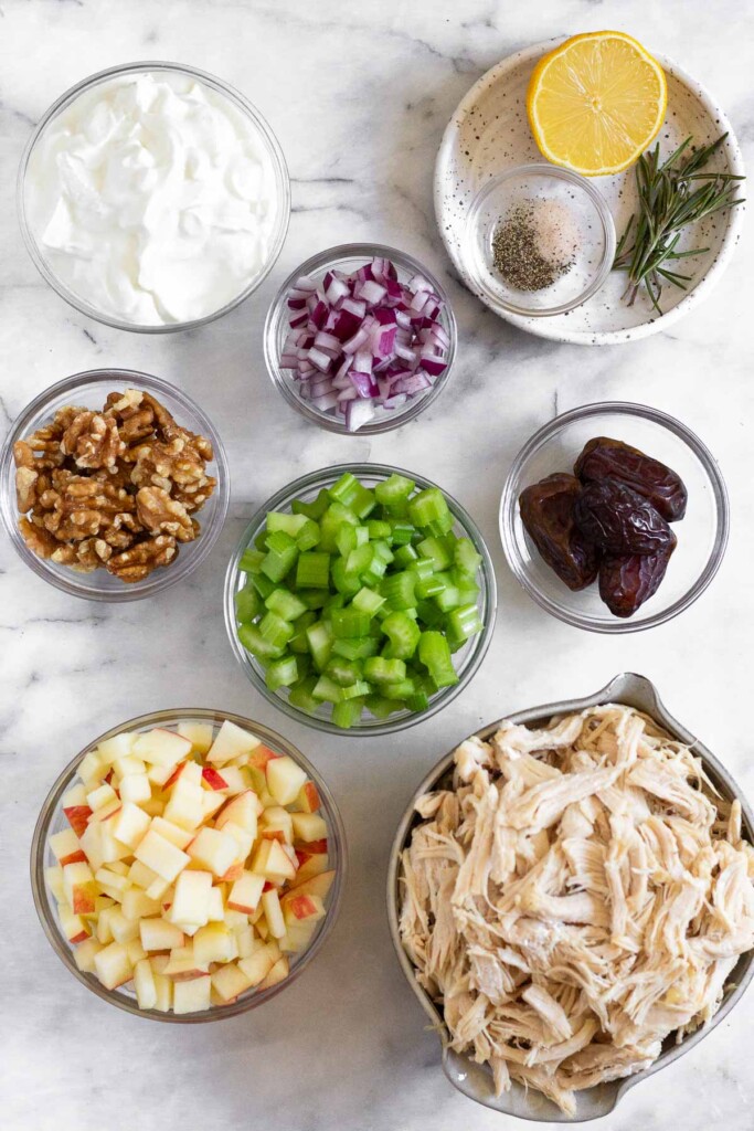 White marble counter with a bowl of diced red onion, a bowl of medjool dates, a bowl of shredded chicken, a bowl of diced apples, a bowl of diced celery, a bowl of walnuts, a bowl of greek yogurt, and a small plate with a bowl of salt and pepper, half a lemon, and fresh rosemary sprigs on it.