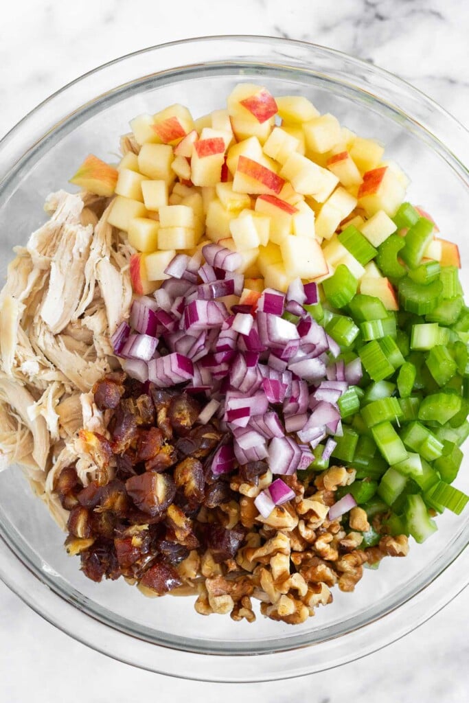 A large glass bowl of shredded chicken, diced apples, diced celery, chopped walnuts, chopped dates, and diced red onion in it before it is all mixed together.