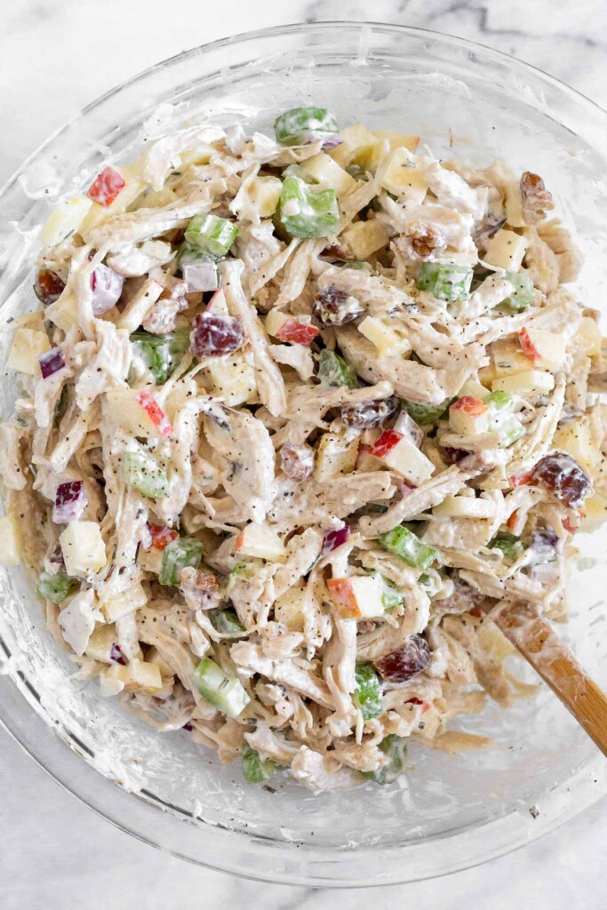 Chicken salad with apples, walnuts, celery, and dates in a large glass bowl with a spatula in it.