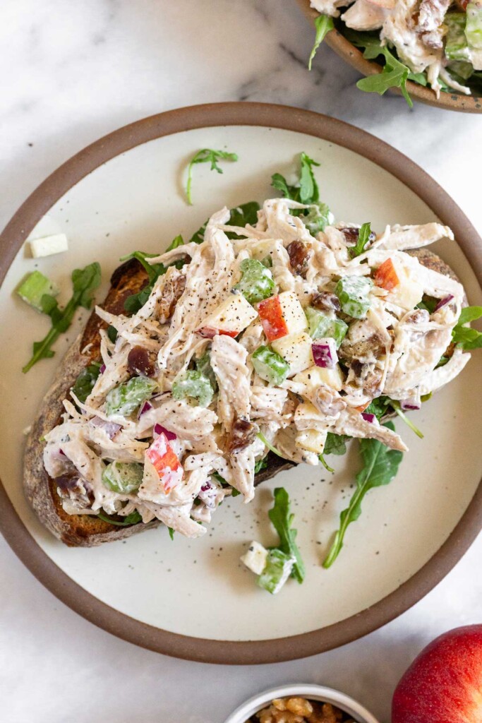 Apple walnut chicken salad on top of arugula on a piece of toast on a plate. Around the plate is a bowl of chicken salad, a red apple, and a ramekin of walnuts.