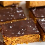 Peanut butter crunch bars topped with chocolate and flaky sea salt lined up on a piece of white parchment paper.