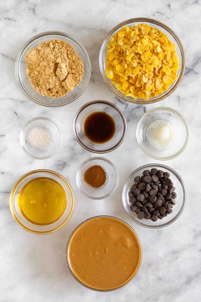 White marble counter with a bowl of cornflakes, a bowl of coconut oil, a bowl of chocolate chips, a bowl of peanut butter, a bowl of honey, a bowl of cinnamon, a bowl of vanilla extract, a bowl of salt, and a bowl of peanut butter powder.