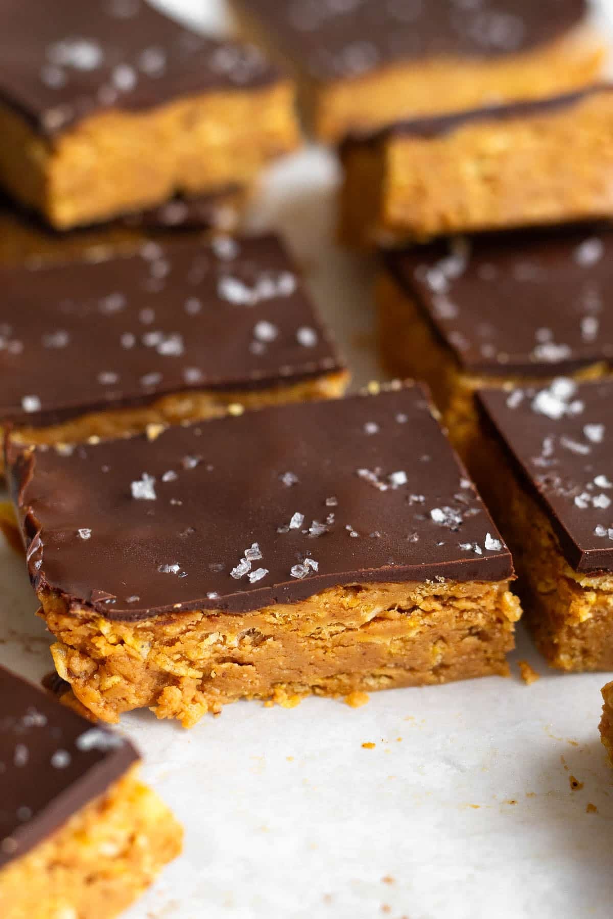 Peanut butter crunch bars topped with chocolate and flaky sea salt lined up on a piece of white parchment paper.