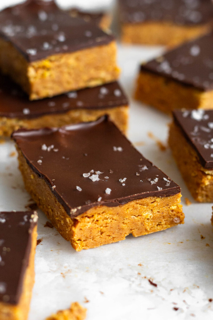 A close up of a chocolate peanut butter crunch bar topped with flaky salt. Around it is more bars with two stacked on top of each other in the back.
