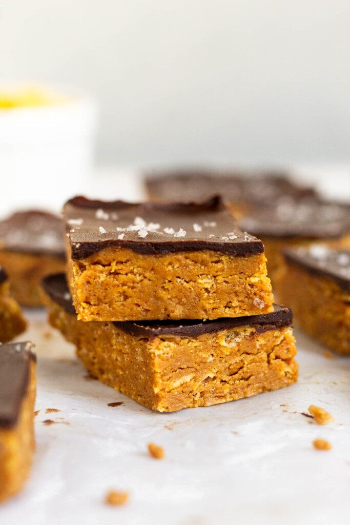 Two chocolate peanut butter crunch bars stacked on top of each other. Around them are more bars and in the far distance is a bowl of cereal.