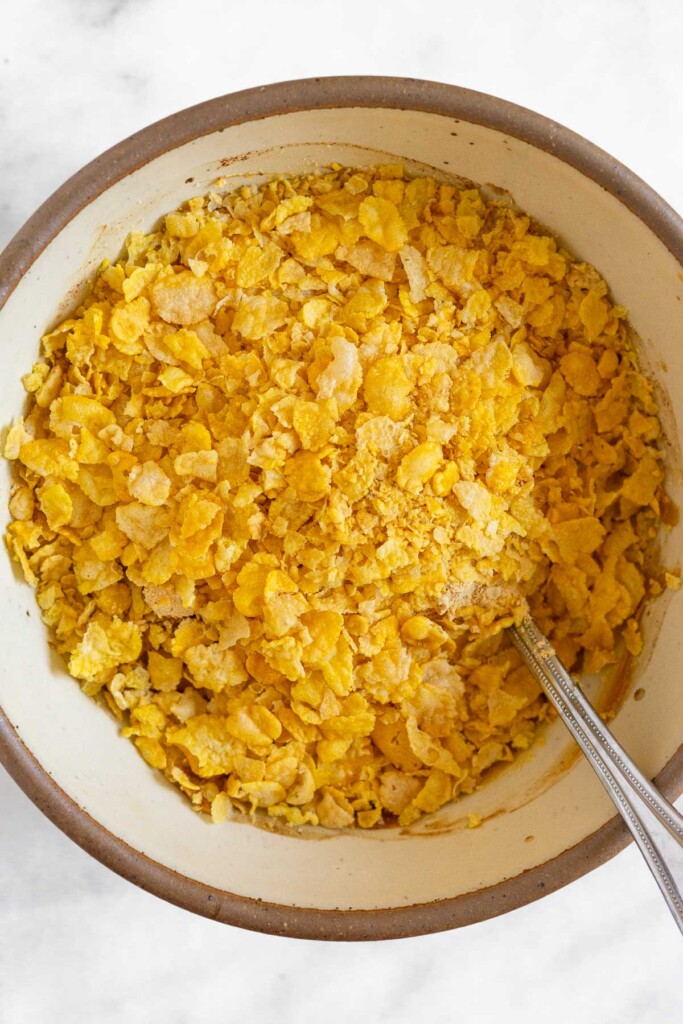 Cornflakes overtop a peanut butter mixture in a white bowl with a spoon in it.