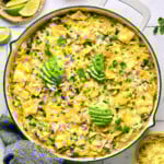 Green chicken enchilada skillet with beans and veggies covered with melted cheese in a large white pan. It is garnished with sliced avocado, diced red onions, and cilantro. The handle of the pan is wrapped in a blue towel and around the pan is a plate of lime wedges, a bowl of diced red onion, cilantro leaves, and a bowl of shredded cheese.
