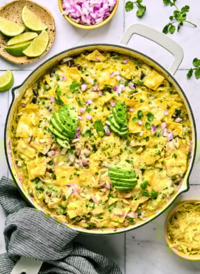 Green chicken enchilada skillet with beans and veggies covered with melted cheese in a large white pan. It is garnished with sliced avocado, diced red onions, and cilantro. The handle of the pan is wrapped in a blue towel and around the pan is a plate of lime wedges, a bowl of diced red onion, cilantro leaves, and a bowl of shredded cheese.