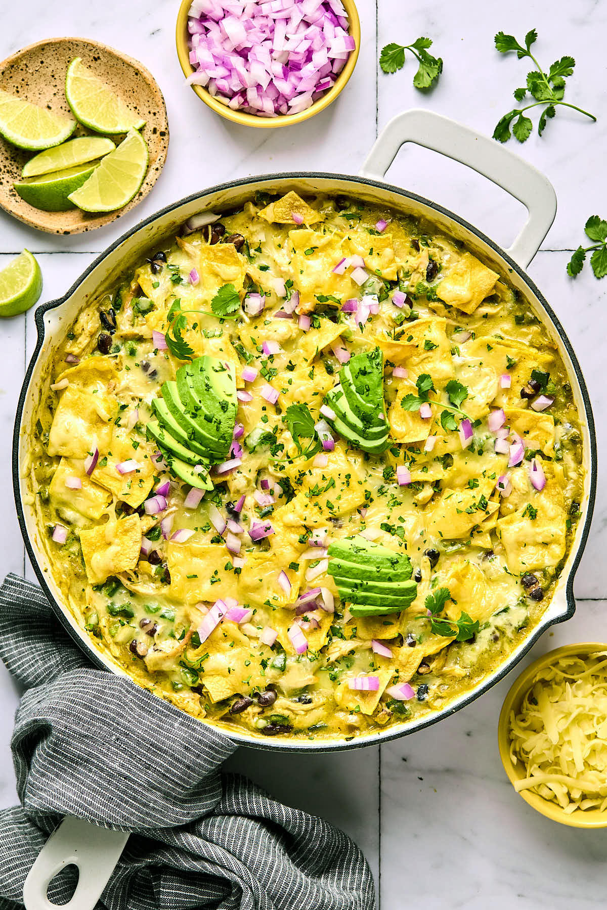 Green chicken enchilada skillet with beans and veggies covered with melted cheese in a large white pan. It is garnished with sliced avocado, diced red onions, and cilantro. The handle of the pan is wrapped in a blue towel and around the pan is a plate of lime wedges, a bowl of diced red onion, cilantro leaves, and a bowl of shredded cheese.