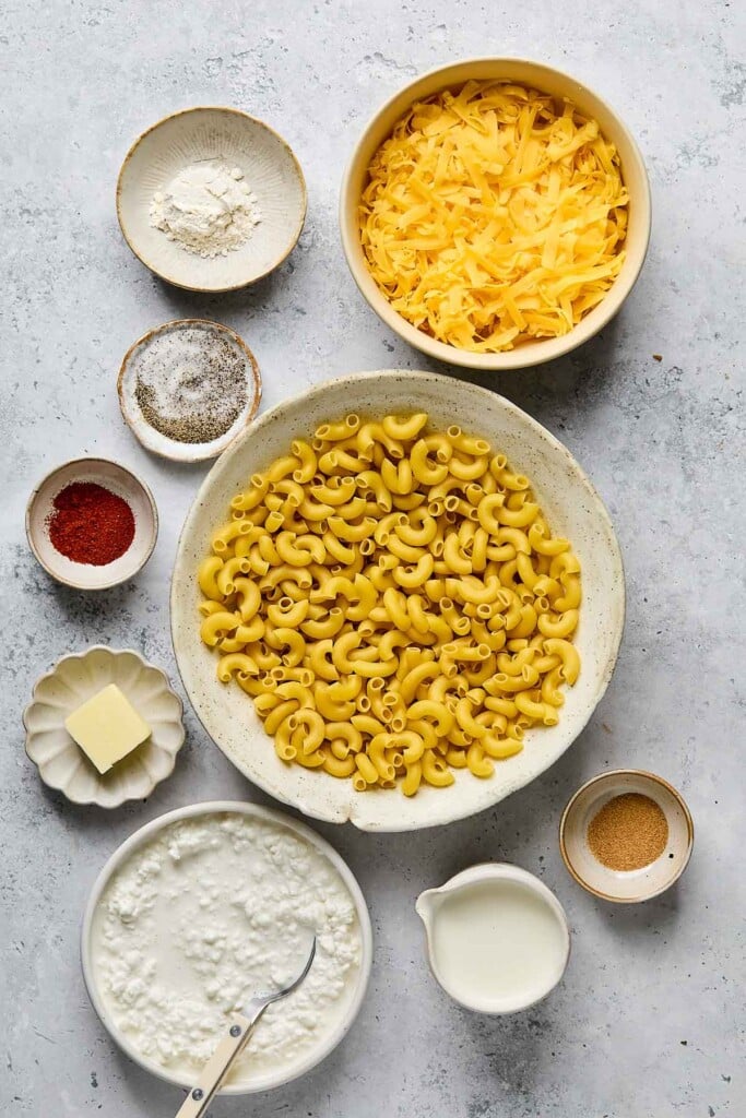 Grey concrete counter with a bowl of shredded cheese, a bowl of dry elbow pasta, a small bowl of spices, a cup of milk, a bowl of cottage cheese with a spoon in it, a small plate of a slice of butter, a bowl of spices, a small bowl of salt and pepper, and a bowl of flour.