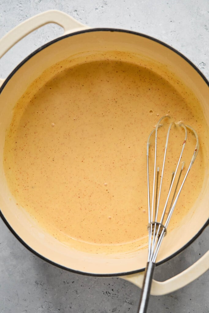 Healthy mac and cheese sauce in a large white dutch oven. There is a metal whisk in the pot as well.