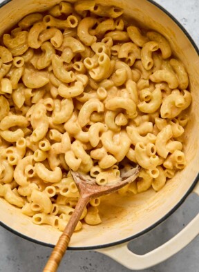 Protein mac and cheese in a large white dutch oven with a wooden spoon in it.