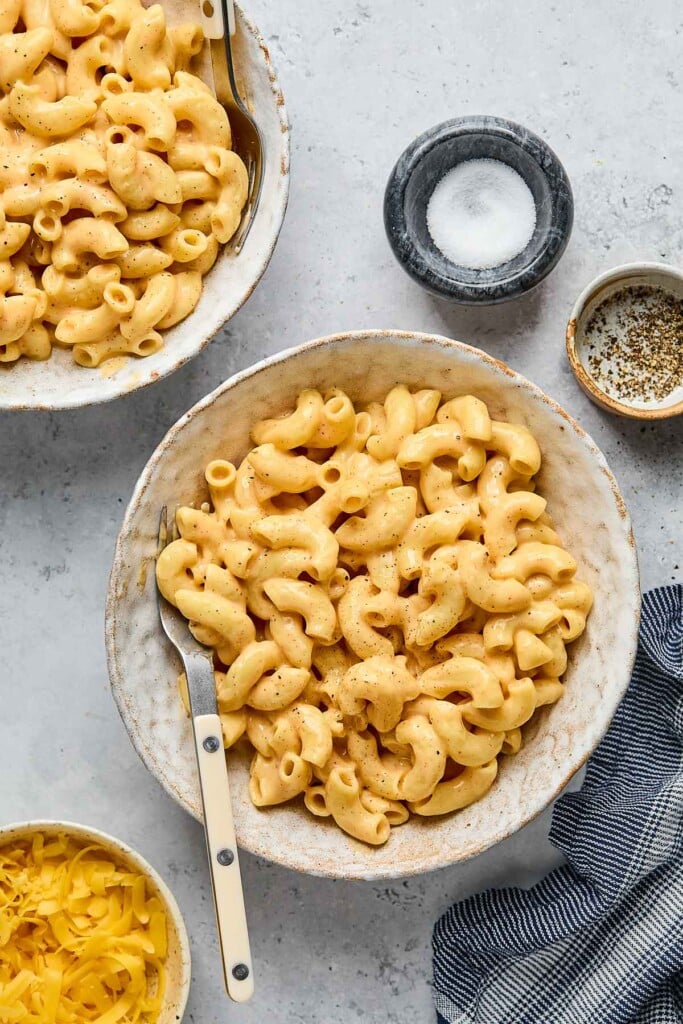 Overhead shot of two bowls of cottage cheese mac and cheese. In both of the bowls is a fork. Around the bowls is a bowl of shredded cheese, a small bowl of salt, a small bowl of pepper, and a blue linen.