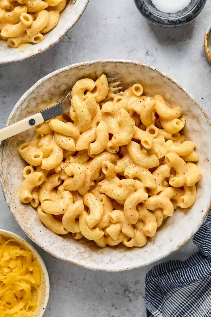 High protein mac and cheese in a white bowl with a fork in it. Around the bowl is a bowl of shredded cheese, another bowl of mac and cheese, a small dish of salt, and a blue linen.