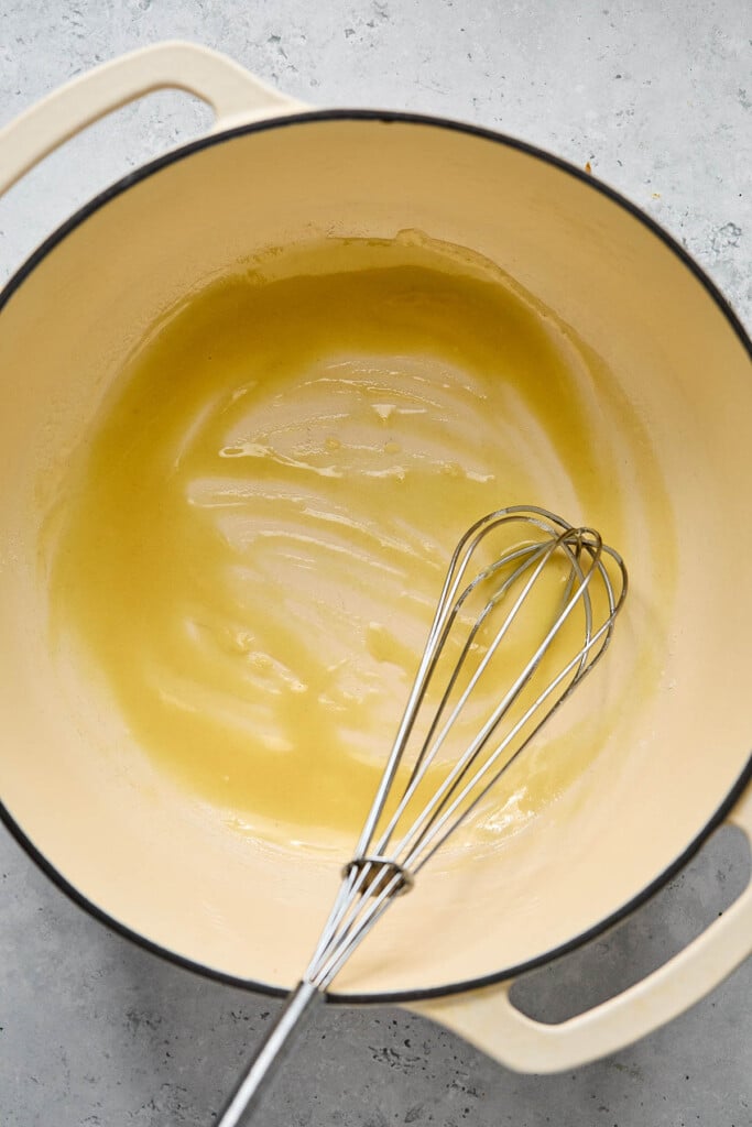 A roux in a large white dutch oven. A metal whisk is in the pot as well.