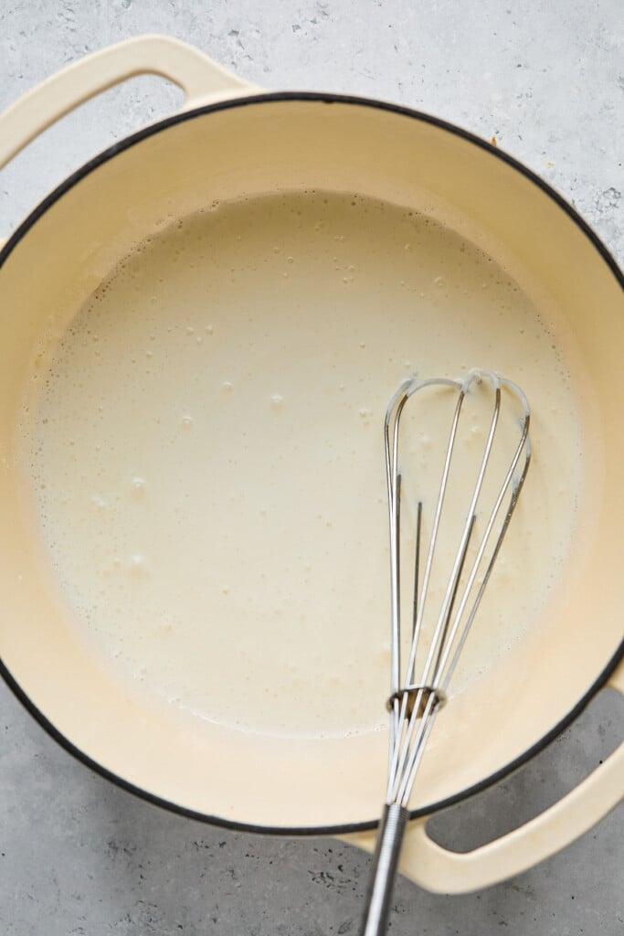 Cottage cheese and milk mixture mixed with a roux in a large white dutch oven. In the pot is a metal whisk.
