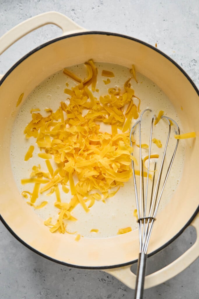Cottage cheese and milk mixture with shredded orange cheese sprinkled on top of it in large white dutch oven. A metal whisk is also in the pan.