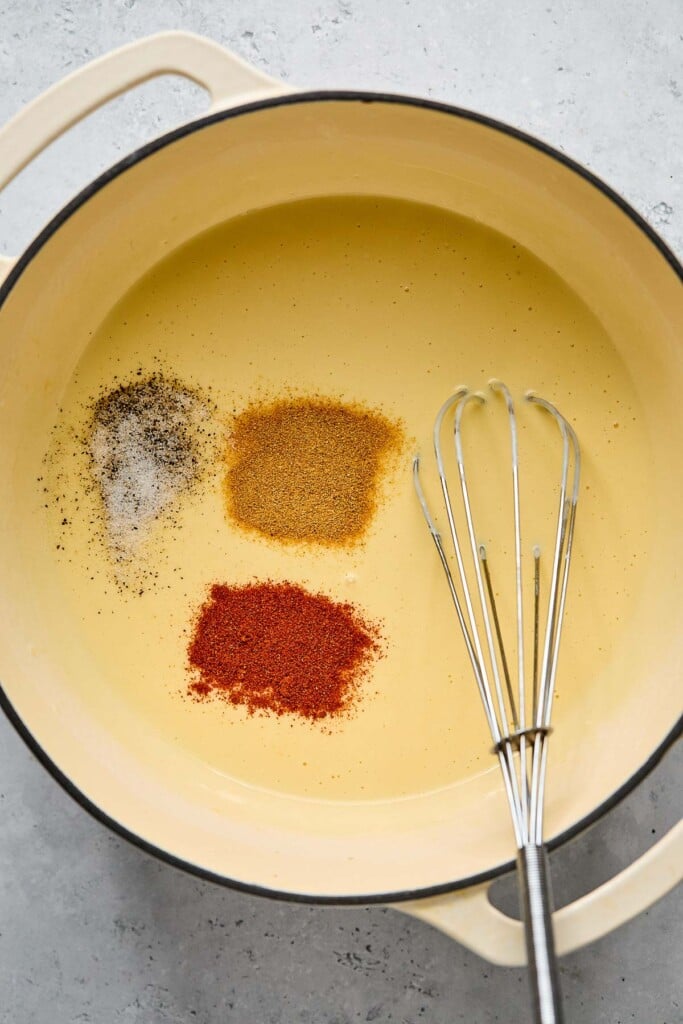 Cheese sauce with salt, pepper, and two different spices sprinkled on top of it in a large white dutch oven. A metal whisk is also in the pan.