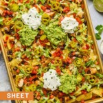 Sheet pan nachos with diced chicken, black beans, cheese, lettuce, tomato, sour cream, guacamole, pickled jalapeños, and cilantro. Next to the sheet pan is a lime that has been cut in half, a bowl of pickled jalapeños, a striped kitchen towel, and cilantro leaves.