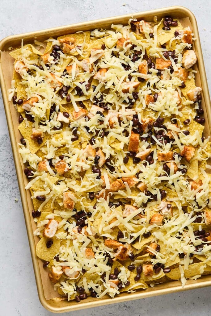 A baking sheet lined with parchment paper layered with tortilla chips, diced chicken, black beans, and shredded cheese.