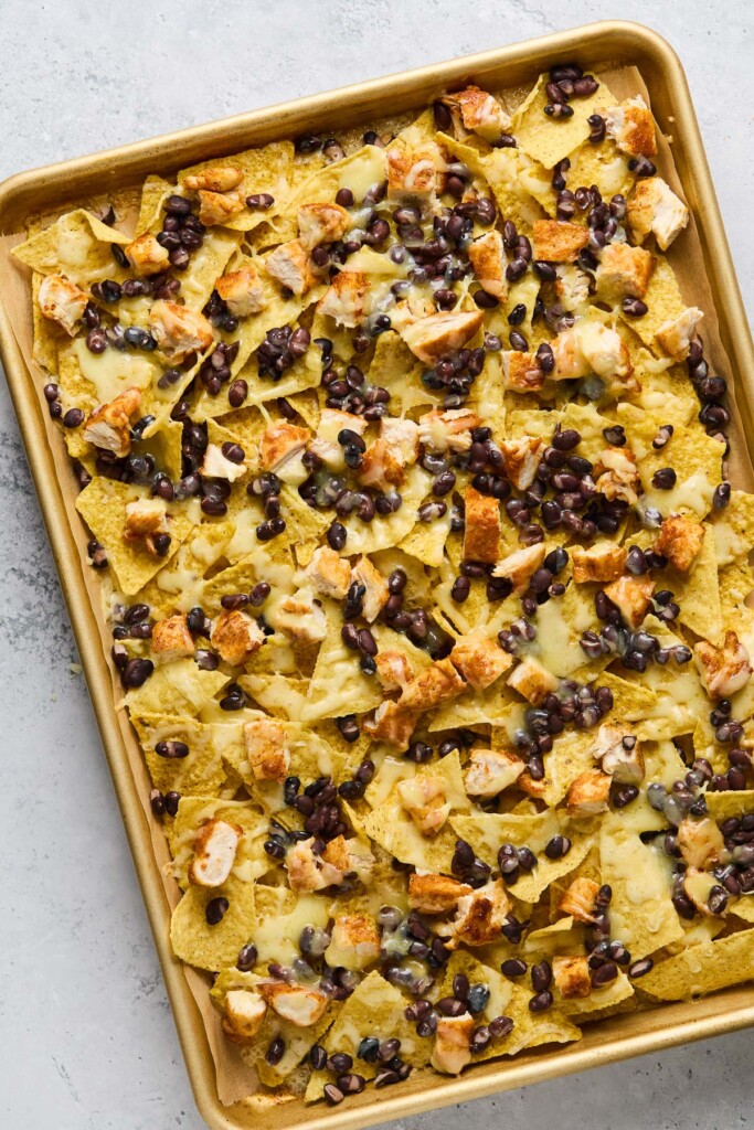 A baking sheet lined with parchment paper layered with tortilla chips, diced chicken, black beans, and melted cheese.
