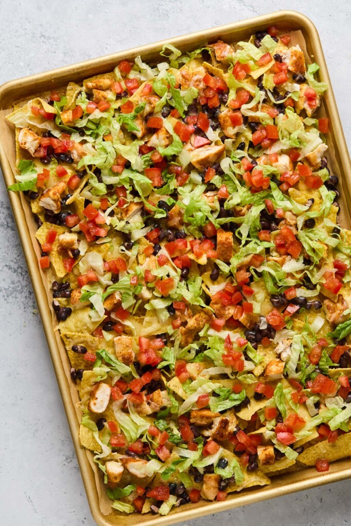 A baking sheet lined with parchment paper layered with tortilla chips, diced chicken, black beans, melted cheese, shredded lettuce, and diced tomaotes.