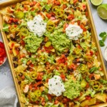 Sheet pan nachos with diced chicken, black beans, cheese, lettuce, tomato, sour cream, guacamole, pickled jalapeños, and cilantro. Next to the sheet pan is a lime that has been cut in half, a bowl of pickled jalapeños, a striped kitchen towel, a bowl of diced tomatoes, cilantro leaves, and a bowl of shredded cheese.