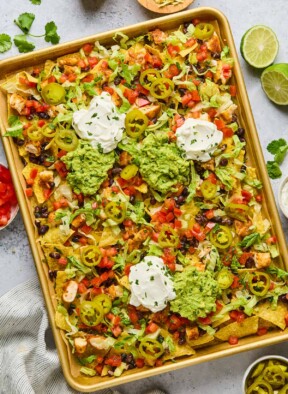 Sheet pan nachos with diced chicken, black beans, cheese, lettuce, tomato, sour cream, guacamole, pickled jalapeños, and cilantro. Next to the sheet pan is a lime that has been cut in half, a bowl of pickled jalapeños, a striped kitchen towel, a bowl of diced tomatoes, cilantro leaves, and a bowl of shredded cheese.
