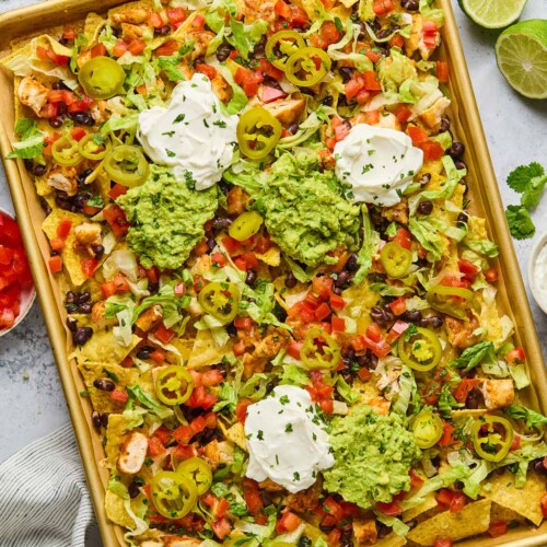 Sheet pan nachos with diced chicken, black beans, cheese, lettuce, tomato, sour cream, guacamole, pickled jalapeños, and cilantro. Next to the sheet pan is a lime that has been cut in half, a bowl of pickled jalapeños, a striped kitchen towel, a bowl of diced tomatoes, cilantro leaves, and a bowl of shredded cheese.