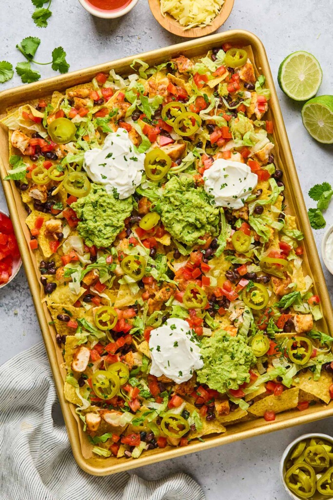 Sheet pan nachos with diced chicken, black beans, cheese, lettuce, tomato, sour cream, guacamole, pickled jalapeños, and cilantro. Next to the sheet pan is a lime that has been cut in half, a bowl of pickled jalapeños, a striped kitchen towel, a bowl of diced tomatoes, cilantro leaves, and a bowl of shredded cheese.