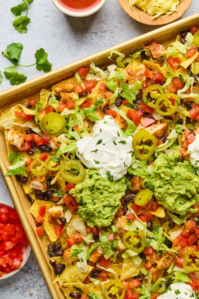 Sheet pan chicken nachos with black beans, cheese, lettuce, tomato, sour cream, guacamole, pickled jalapeños, and cilantro. Around the sheet pan is a bowl of diced tomatoes, some cilantro leaves, a bowl of salsa, and a bowl of shredded cheese.