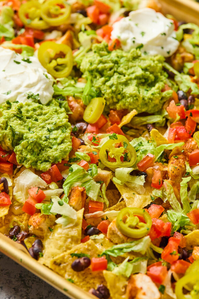 A close up of sheet pan nachos layered with tortilla chips, chicken, black beans, lettuce, tomatoes, pickled jalapeños, sour cream, guacamole, and cilantro.
