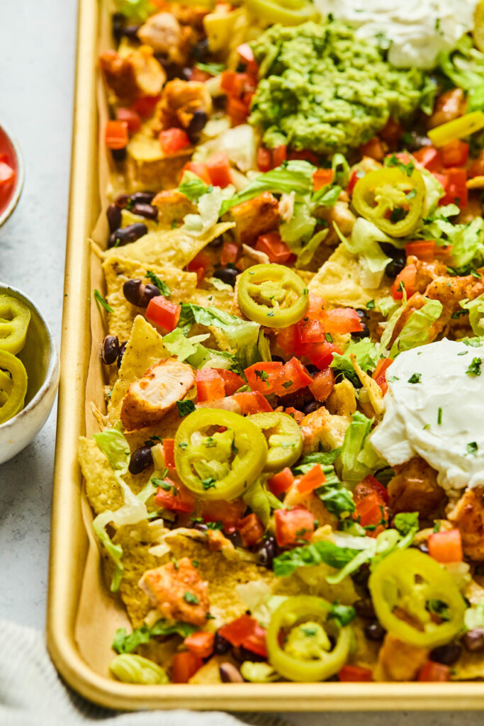 Sheet pan nachos with diced chicken, black beans, melted cheese, lettuce, tomatoes, pickled jalapeños, sour cream, and guacamole. It is garnished with cilantro. Next to the pan off to the side is a bowl of bowl of pickled jalapeños.