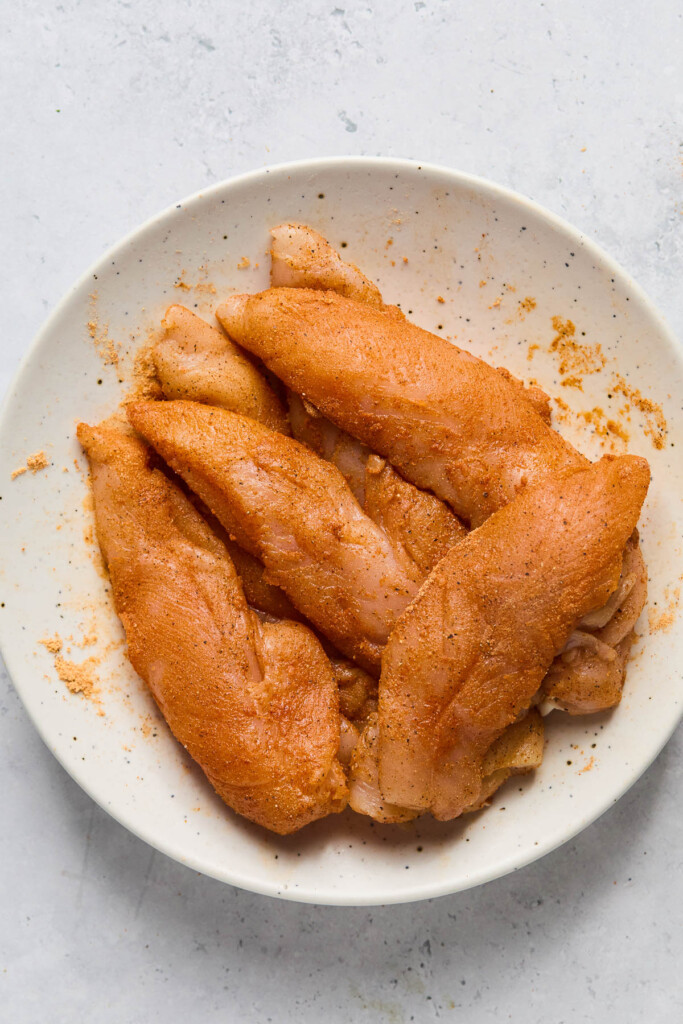 Raw chicken tenders that have been seasoned with taco seasoning in a white bowl.