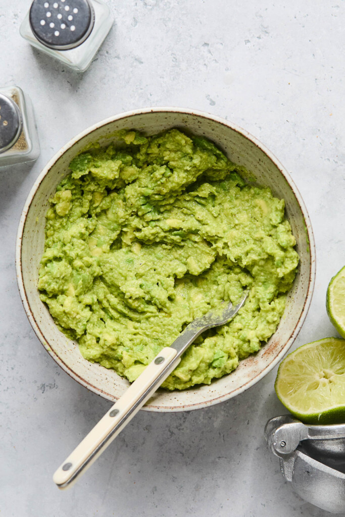 A white bowl filled with guacamole with a fork in it. Around the bowl is a salt and pepper shaker, a lime that has been cut in half, and a silver citrus press.