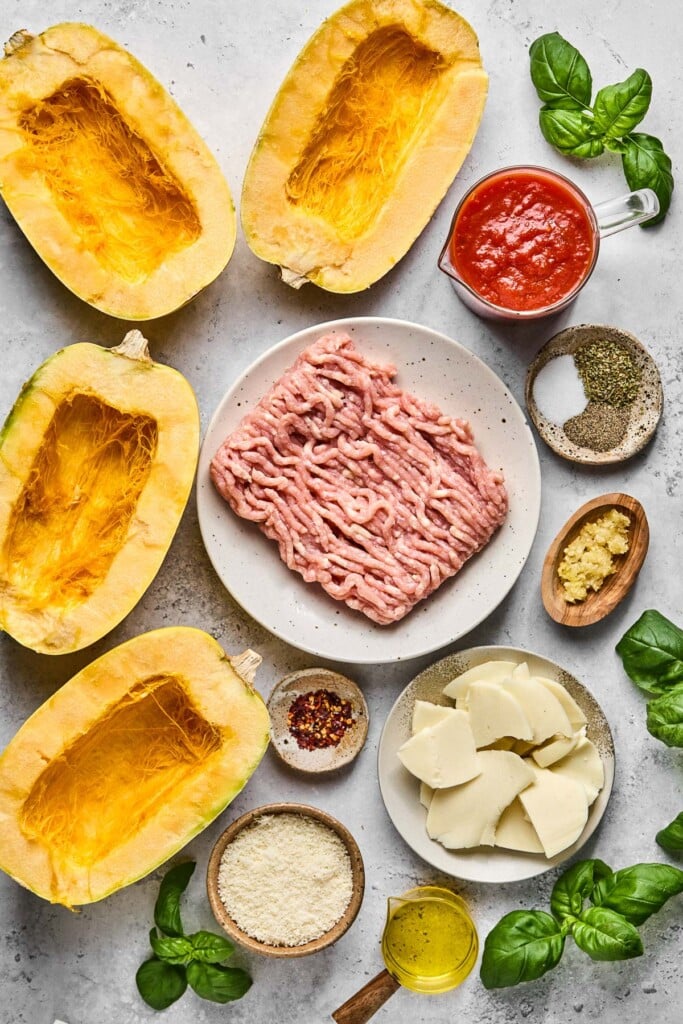 Grey concrete counter with four halves of spaghetti squash, a plate of raw ground chicken, a measuring cup of marinara sauce, a small plate of spices, a small dish of minced garlic, a plate of pieces of sliced cheese, leaves of fresh basil, a small dish of olive oil, a bowl of grated cheese, and a small bowl of red pepper flakes.