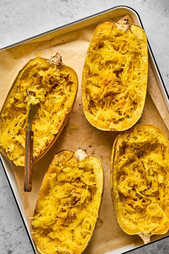 Baking sheet lined with parchment paper with four halves of spaghetti squash that have been baked and threaded. A fork is in one of them.