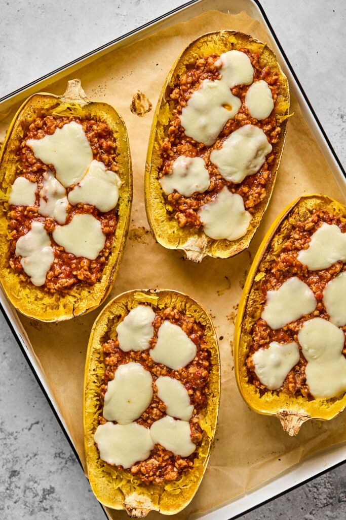 Four cooked spaghetti squash halves filled with meat sauce and topped with melted cheese on a parchment paper lined baking sheet.