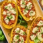 Four chicken parmesan spaghetti squash boats on parchment paper lined baking sheet. They are garnished with red peppers flakes and fresh basil. Around the baking sheet is more fresh basil, a bowl of parmesan cheese, a grey kicthen towel, and a bowl of red pepper flakes.