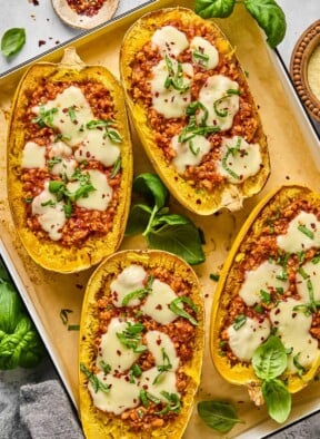 Four chicken parmesan spaghetti squash boats on parchment paper lined baking sheet. They are garnished with red peppers flakes and fresh basil. Around the baking sheet is more fresh basil, a bowl of parmesan cheese, a grey kicthen towel, and a bowl of red pepper flakes.