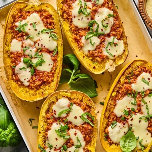 Four chicken parmesan spaghetti squash boats on parchment paper lined baking sheet. They are garnished with red peppers flakes and fresh basil. Around the baking sheet is more fresh basil, a bowl of parmesan cheese, a grey kicthen towel, and a bowl of red pepper flakes.
