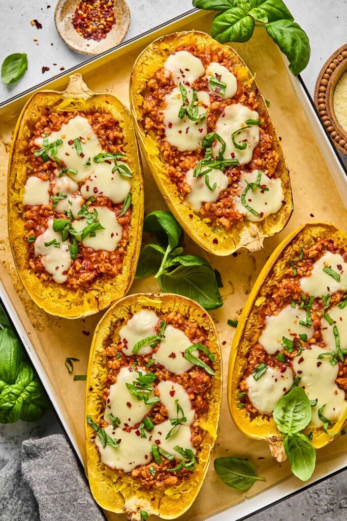 Four chicken parmesan spaghetti squash boats on parchment paper lined baking sheet. They are garnished with red peppers flakes and fresh basil. Around the baking sheet is more fresh basil, a bowl of parmesan cheese, a grey kicthen towel, and a bowl of red pepper flakes.