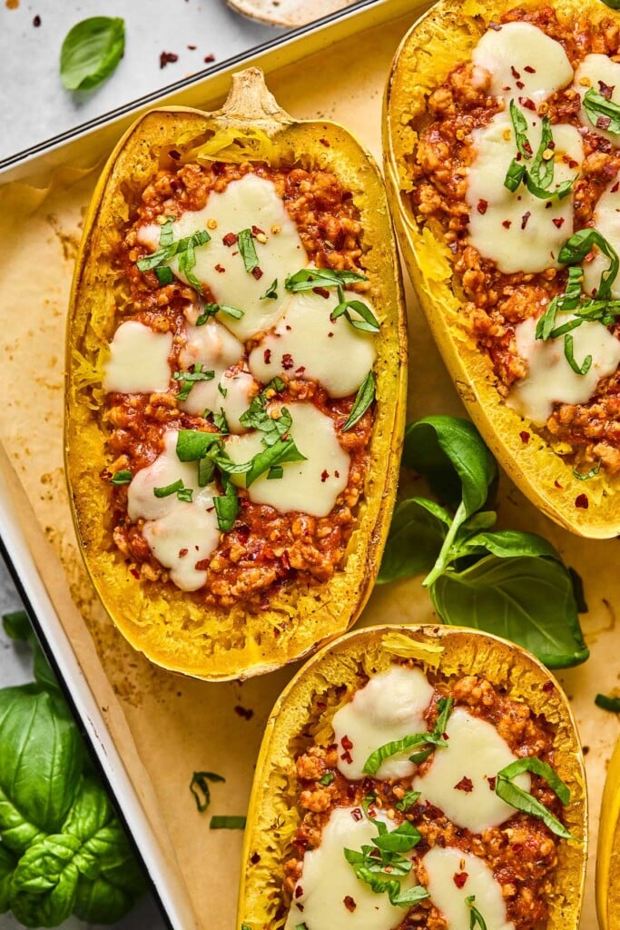 Overhead shot of spaghetti squash chicken parmesan boats topped with melted mozzarella cheese, fresh basil, and red pepper flakes. They are on a baking sheet and around the baking sheet is more fresh basil.