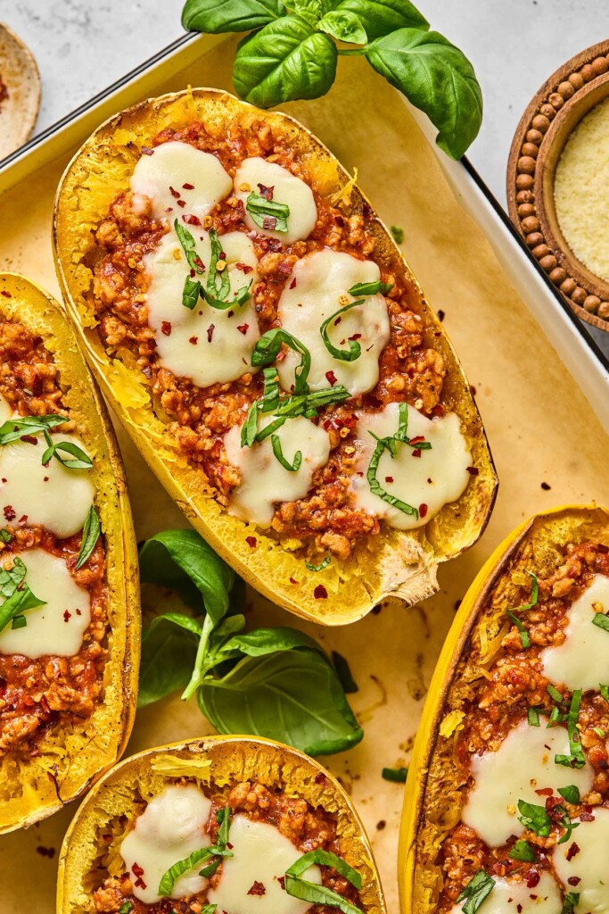 A baking sheet full of spaghetti squash halves stuffed with meat sauce sauce and topped with melted cheese, fresh basil, and red pepper flakes. Around the pan is more fresh basil and a bowl of shredded cheese.