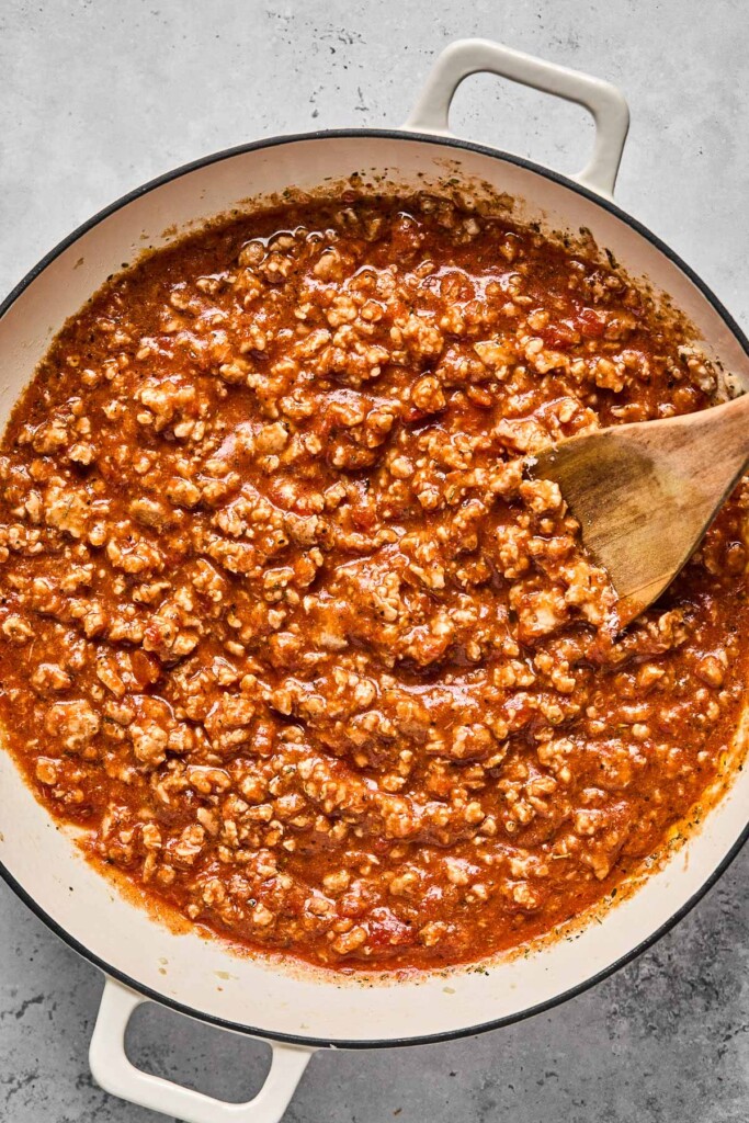 Large white skillet filled with marinara sauce mixed with ground meat. A wooden spoon is also in the pan.
