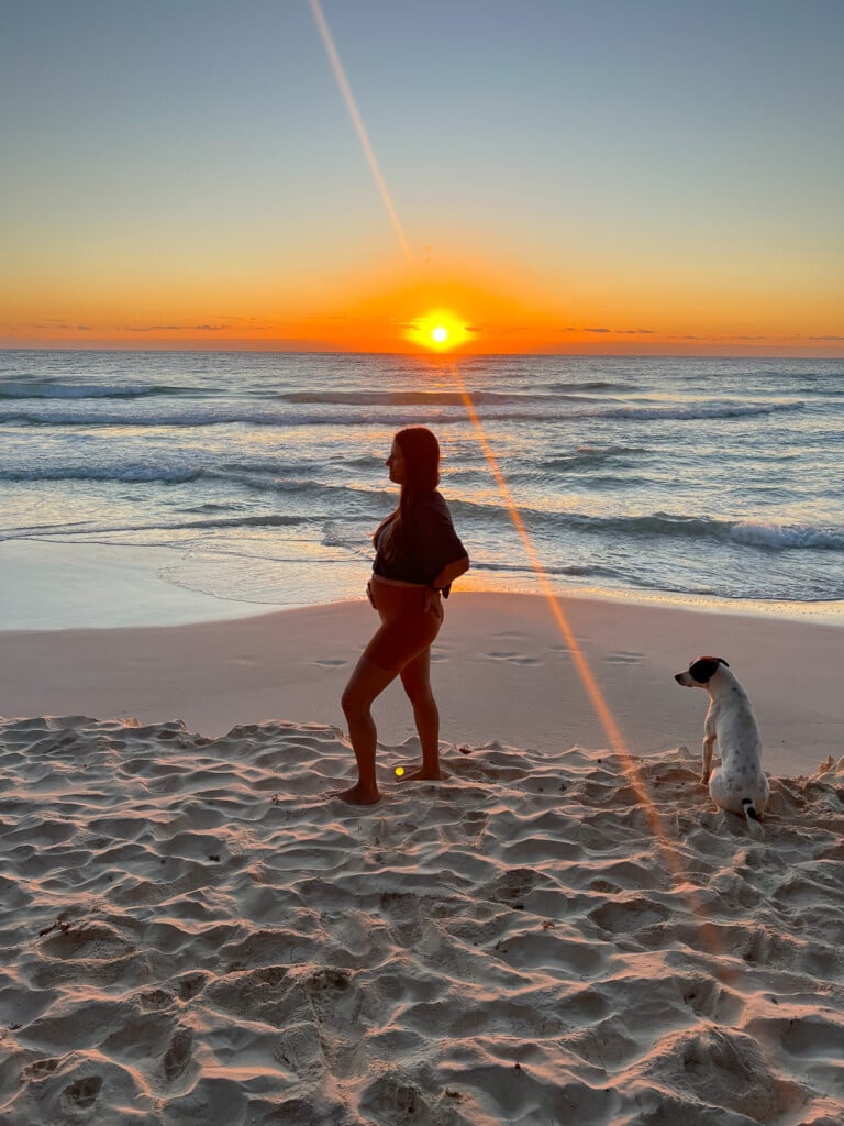 Pregnant woman on the beach with the sun rising behind her. She is standing to show off her bump and next to her is a dog looking at her.