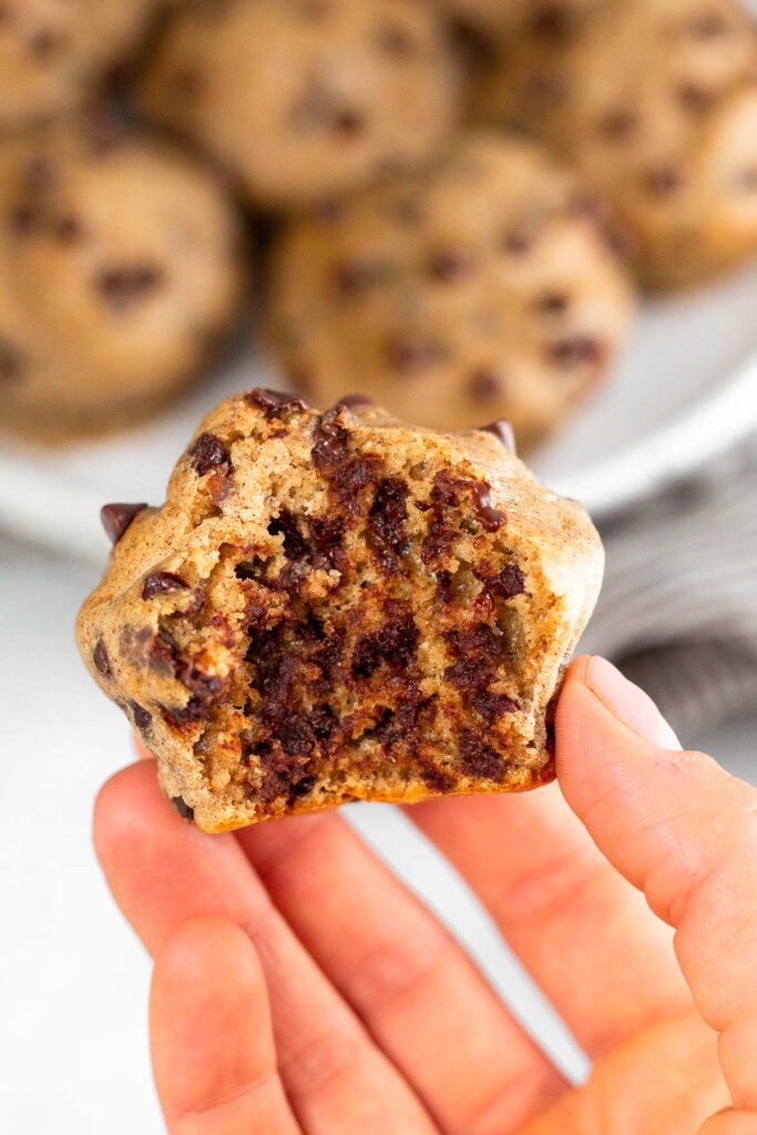 A hand holding a cottage cheese muffin with a bite taken out of it. In the background is a plate of more muffins.