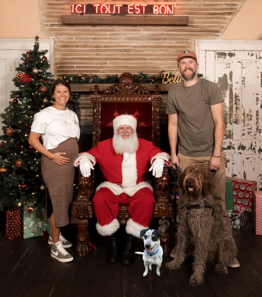 A husband, a pregnant wife, and their two dogs taking a picture with Santa. Around them is a decorated Christmas tree and some wrapped presents.