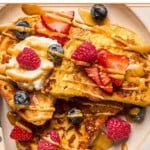 A close up of a plate of protein french toast topped with fresh strawberries, fresh blueberries, fresh raspberries, a dollop of greek yogurt, peanut butter, and maple syrup.