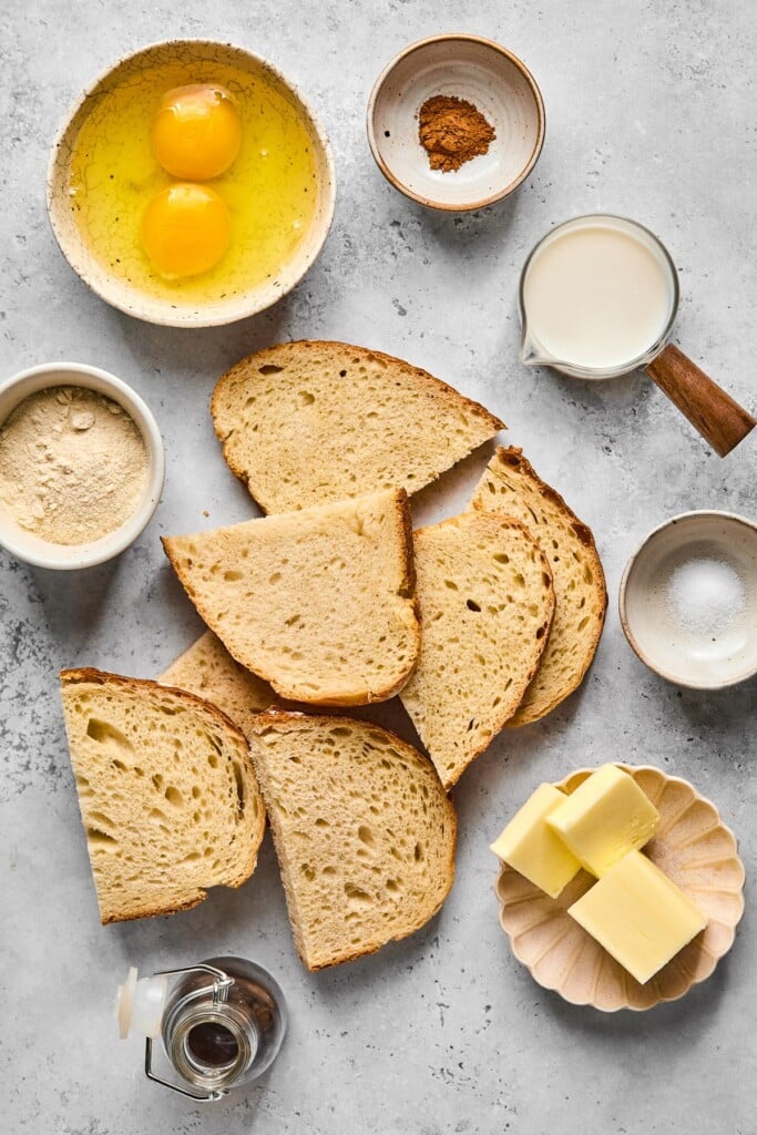 Grey concrete counter with a bowl of cinnamon, a measuring cup of milk, a pile of slices of sourdough bread, a bowl of salt, a plate of slices of butter, a jar of syrup, a bowl of protein powder, and a bowl of two cracked eggs.
